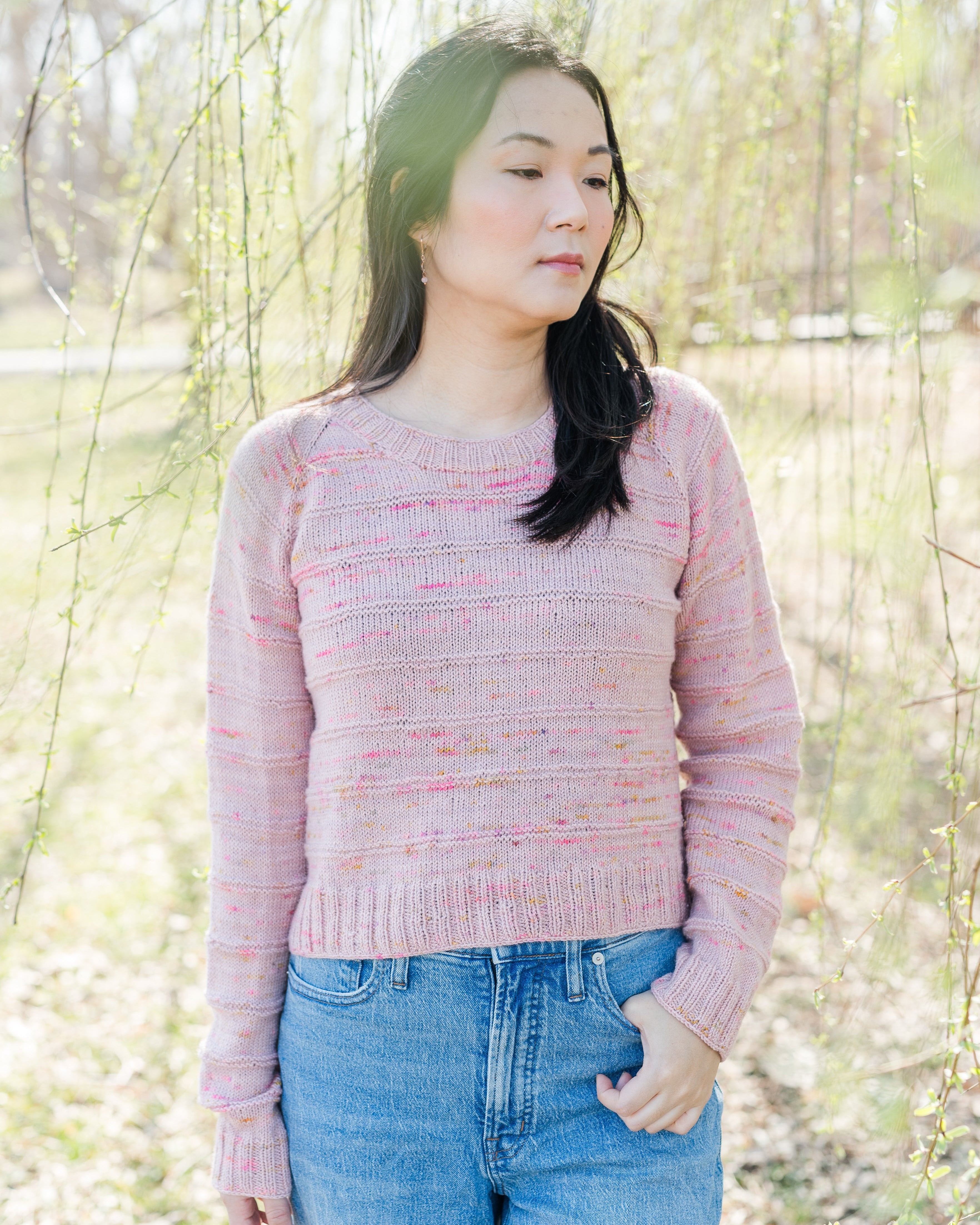 A person wearing a pink knitted Relax Tee with a crew neckline, standing outdoors with trees in the background.