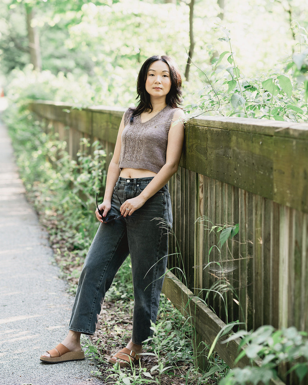 Woman standing outdoors near a wooden fence with greenery in the background